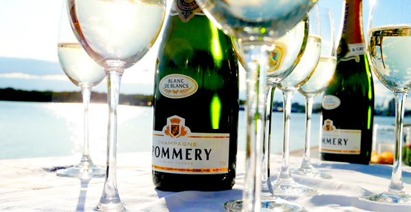 Glas med champagne, flaskor Pommery utomhus vid vattnet i solen.